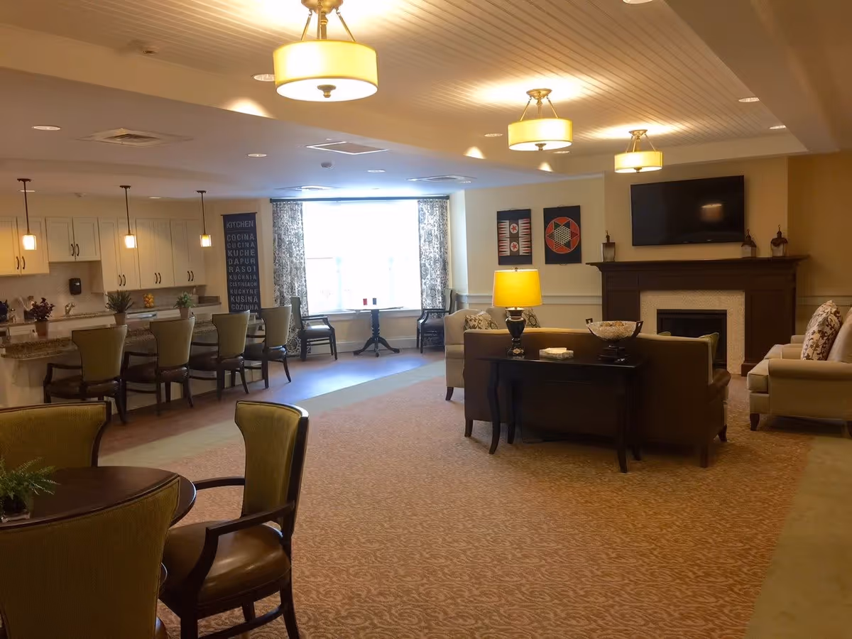 A spacious living area with a seating arrangement including sofas and armchairs around a coffee table and a fireplace with a mounted TV above it. To the left, there is a kitchen counter with bar stools and pendant lights hanging from the ceiling. A large window with patterned curtains allows natural light into the room. The room is decorated with framed artwork on the walls and table lamps providing warm lighting.