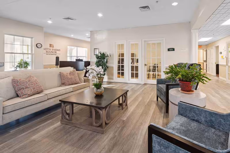 A bright and spacious living room area in Smith Farms Manor featuring a beige sofa with patterned cushions, two gray armchairs, a wooden coffee table with a decorative plant, and a side table with another potted plant. The room has light wood flooring, white walls, and glass-paneled double doors leading to another room. In the background, there is a reception desk and windows letting in natural light.