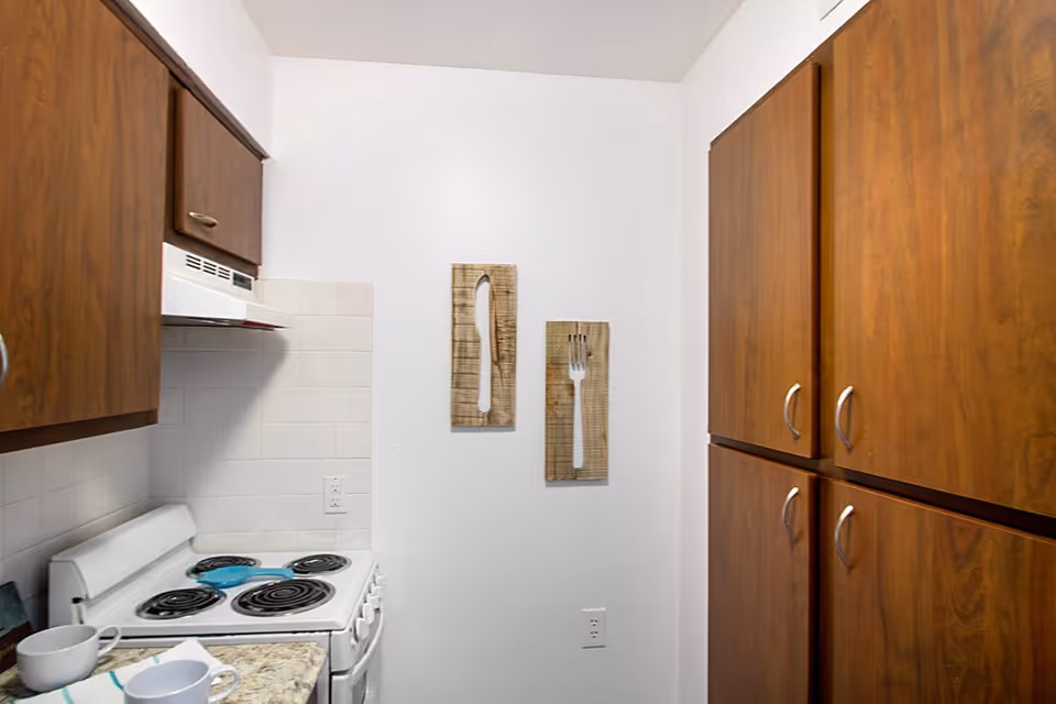 Small kitchen area with a white electric stove and oven, wooden cabinets on both sides, a countertop with two white mugs, and wall decorations shaped like a knife and fork on a white wall.