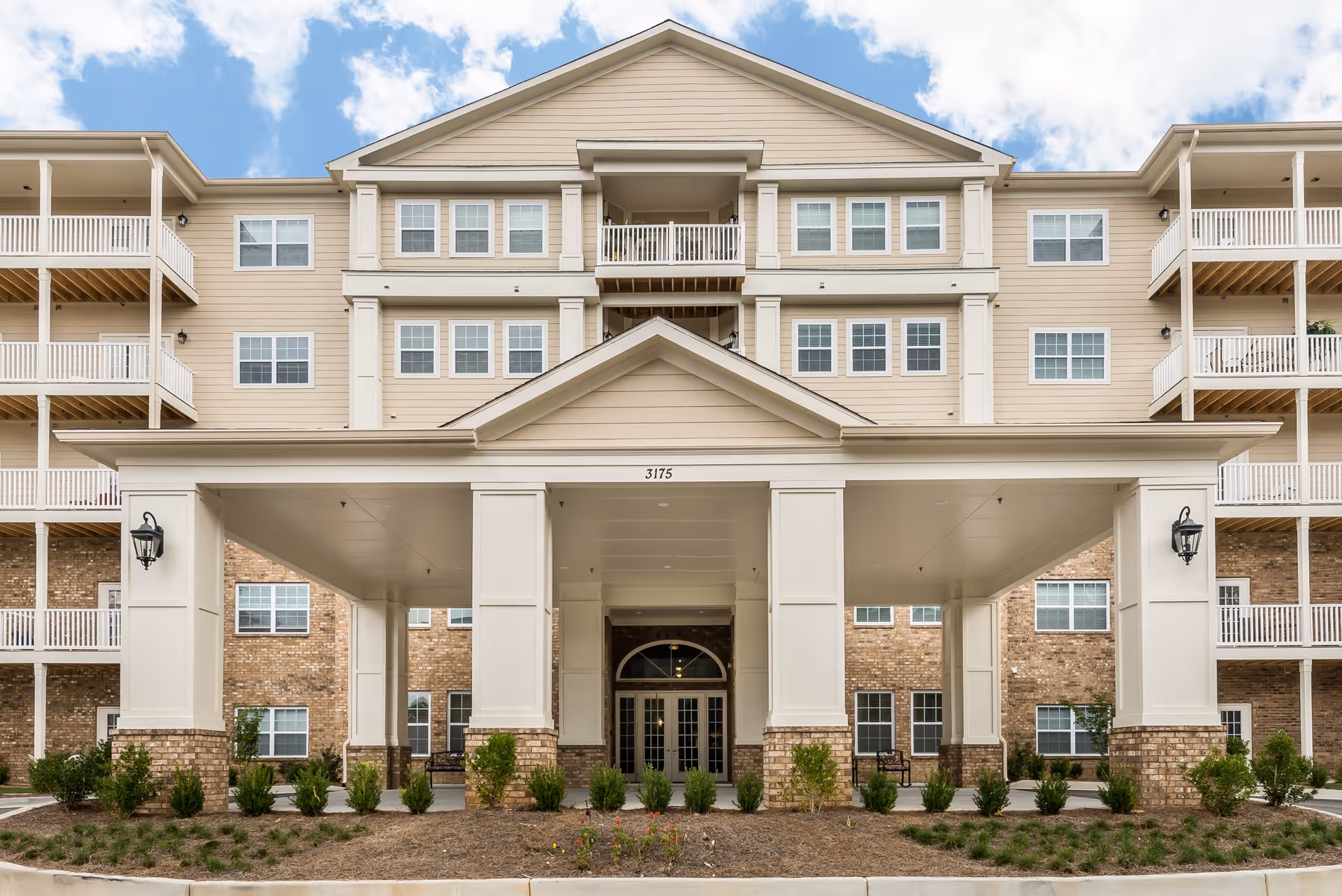 Front exterior view of a multi-story senior living facility building with beige siding, brick accents, multiple windows, balconies, and a covered entrance with columns and outdoor lighting.