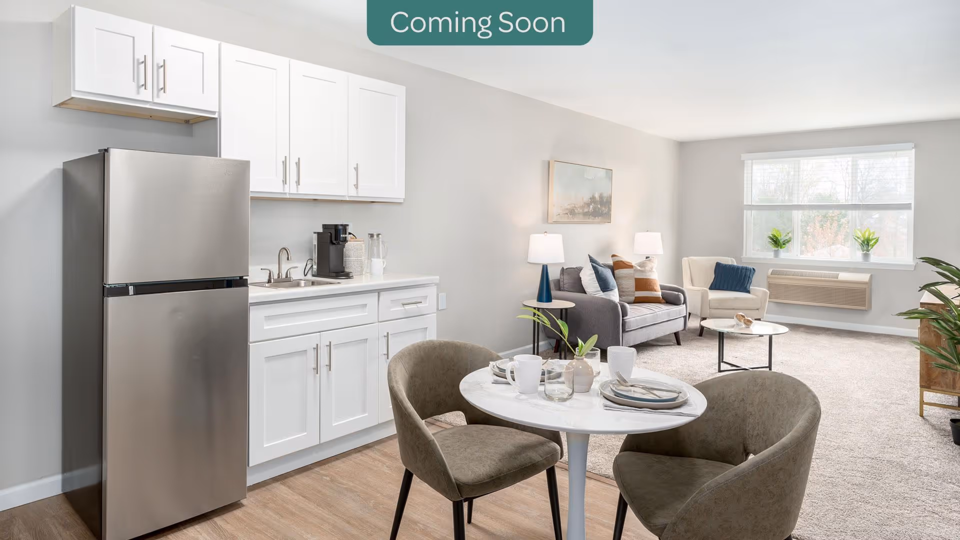 Open living space with a small white kitchenette, round dining table with chairs, and a sofa by a large window.