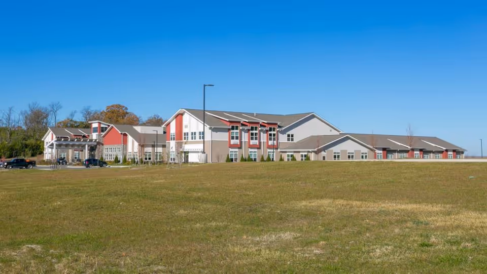 Exterior view of a large, modern senior living facility building with red and beige accents, surrounded by a grassy field under a clear blue sky.