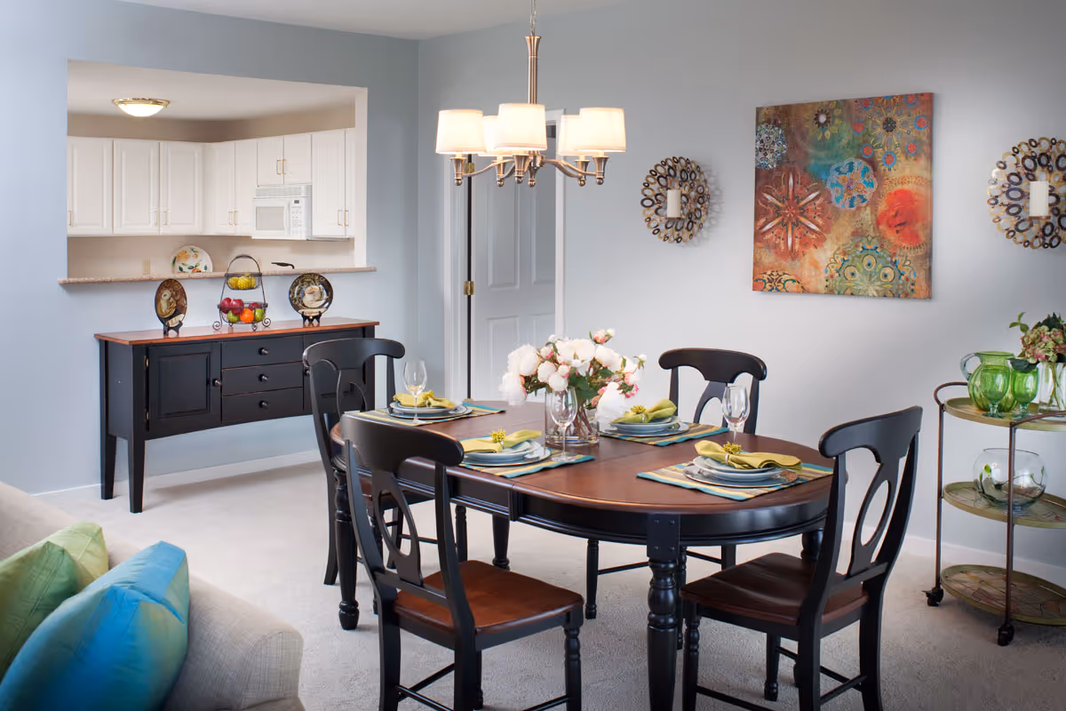 A dining area with a dark wooden oval table set for four with plates, napkins, and glasses. Four matching chairs surround the table. A chandelier with five lights hangs above. Behind the table is a black sideboard with decorative plates and a fruit basket. The kitchen with white cabinets and a microwave is visible through a pass-through window. The walls are light gray with colorful artwork and decorative candle holders. A small round serving cart with green glassware and flowers is on the right side.