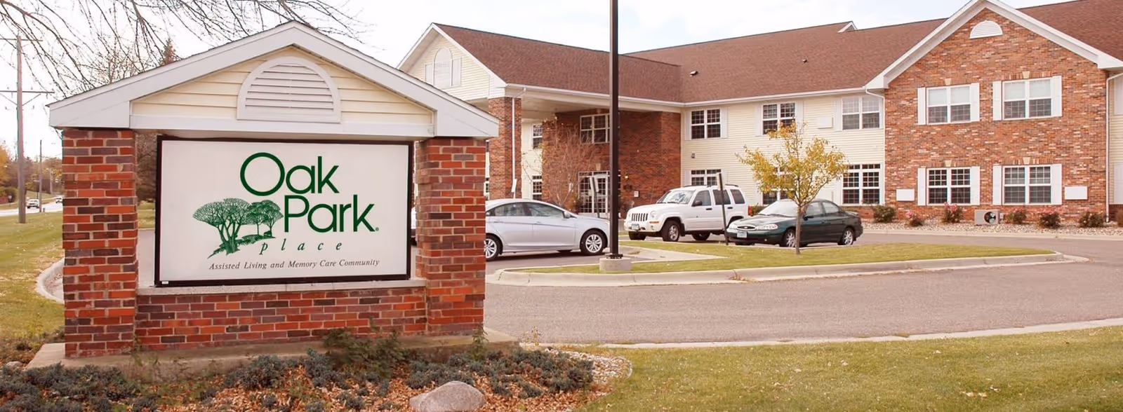 Exterior view of Oak Park Place Albert Lea assisted living and memory care community showing a brick sign with the facility name and a two-story building with parked cars in front.