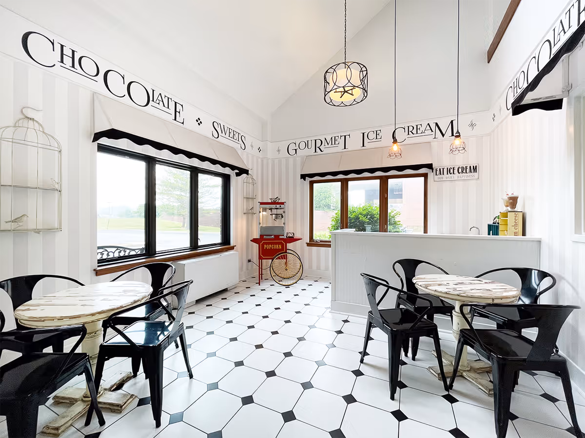 Bright and clean indoor seating area with two round tables surrounded by black metal chairs. The room has white walls with vertical stripes and large windows letting in natural light. Decorative text on the upper walls reads 'CHOCOLATE SWEETS' and 'GOURMET ICE CREAM'. A red popcorn machine is positioned near the windows, and three pendant lights hang from the ceiling.