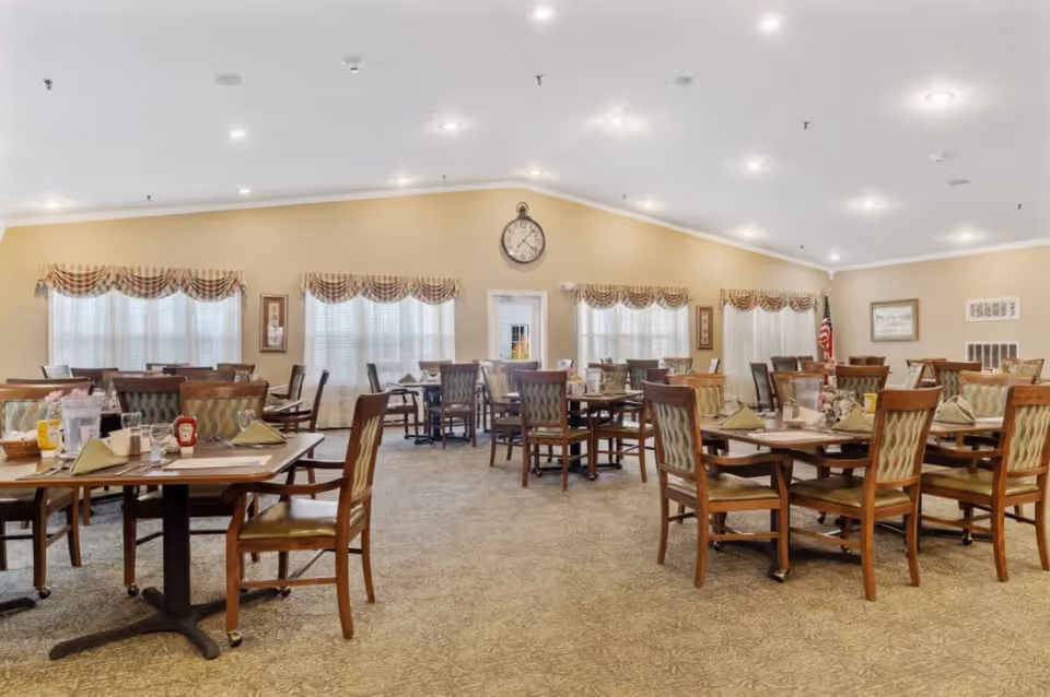 A spacious dining room with multiple wooden tables and chairs arranged neatly. Each table is set with napkins, condiments, and tableware. The room has beige walls, carpeted floors, and windows with patterned valances letting in natural light. A large clock is mounted on the far wall above a door, and an American flag is visible in the corner.