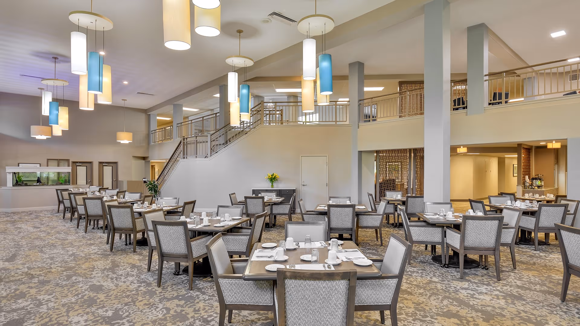 Spacious dining room with multiple tables and chairs arranged neatly, each table set with white cups, plates, and silverware. The room features high ceilings with modern hanging light fixtures in white and blue, a staircase leading to an upper level, and a large fish tank along one wall.