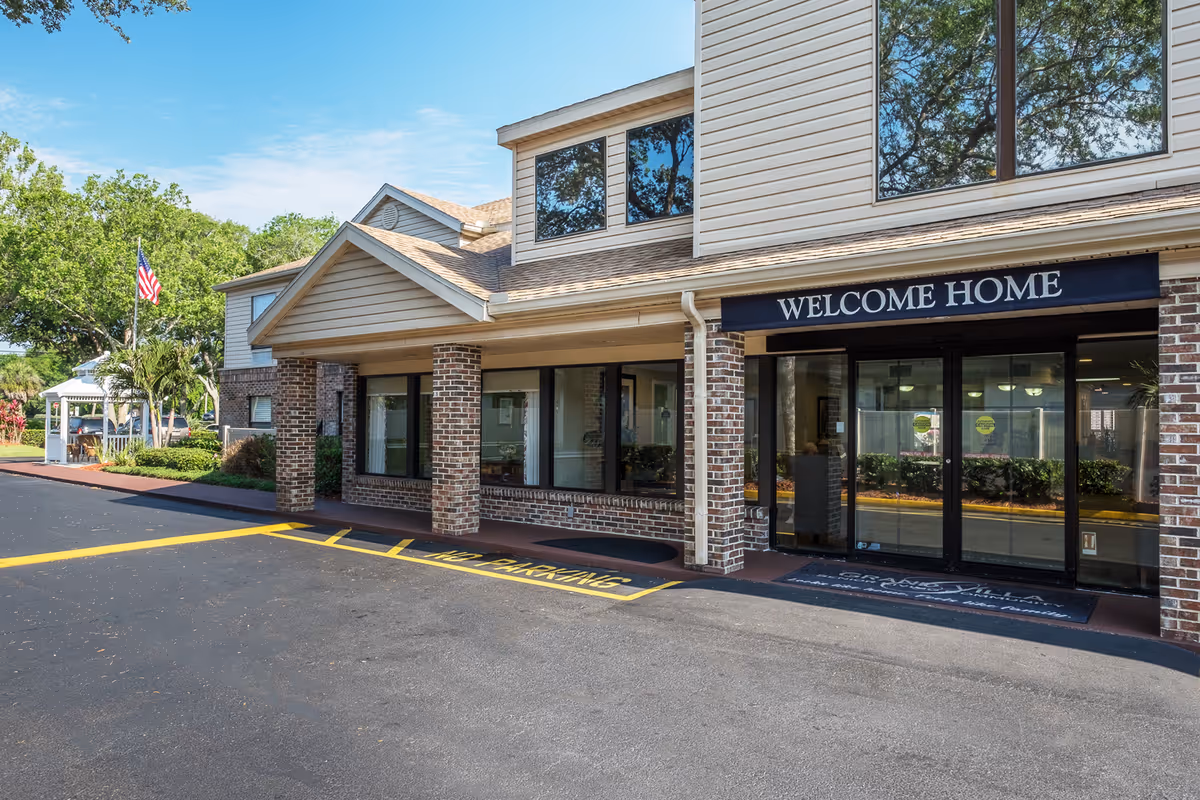 Exterior view of the entrance to Grand Villa of Ormond Beach senior living facility with a 'WELCOME HOME' sign above the glass doors, brick and siding facade, and a parking area with yellow no parking markings.