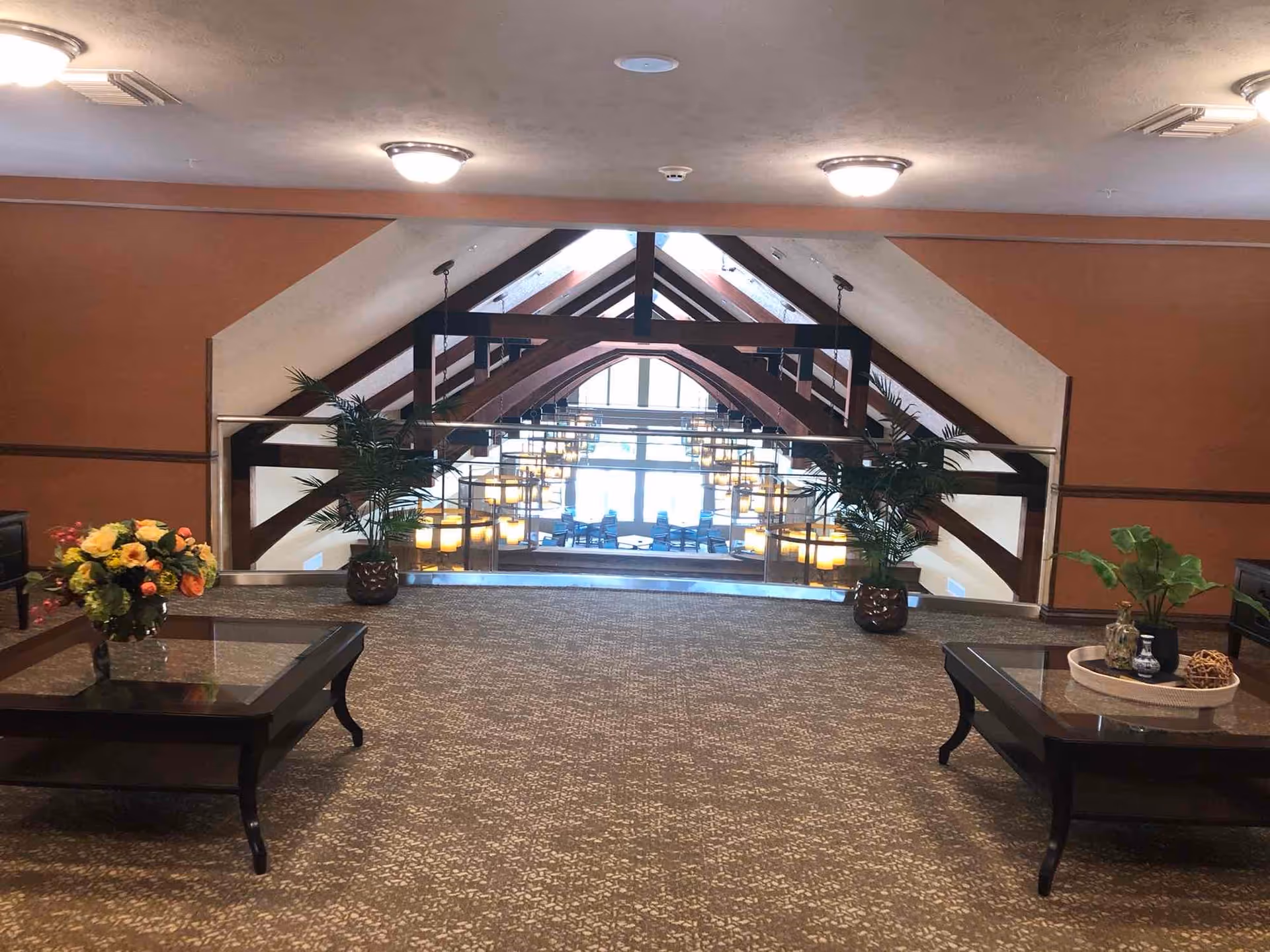 Interior view of a senior living facility lounge area with a patterned carpet, two dark wood coffee tables with glass tops, decorative plants and flowers on the tables, and a large window with wooden beams and hanging lights in the background.