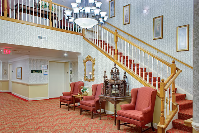 Lobby interior with a grand staircase, chandelier, red upholstered chairs and patterned red carpet.