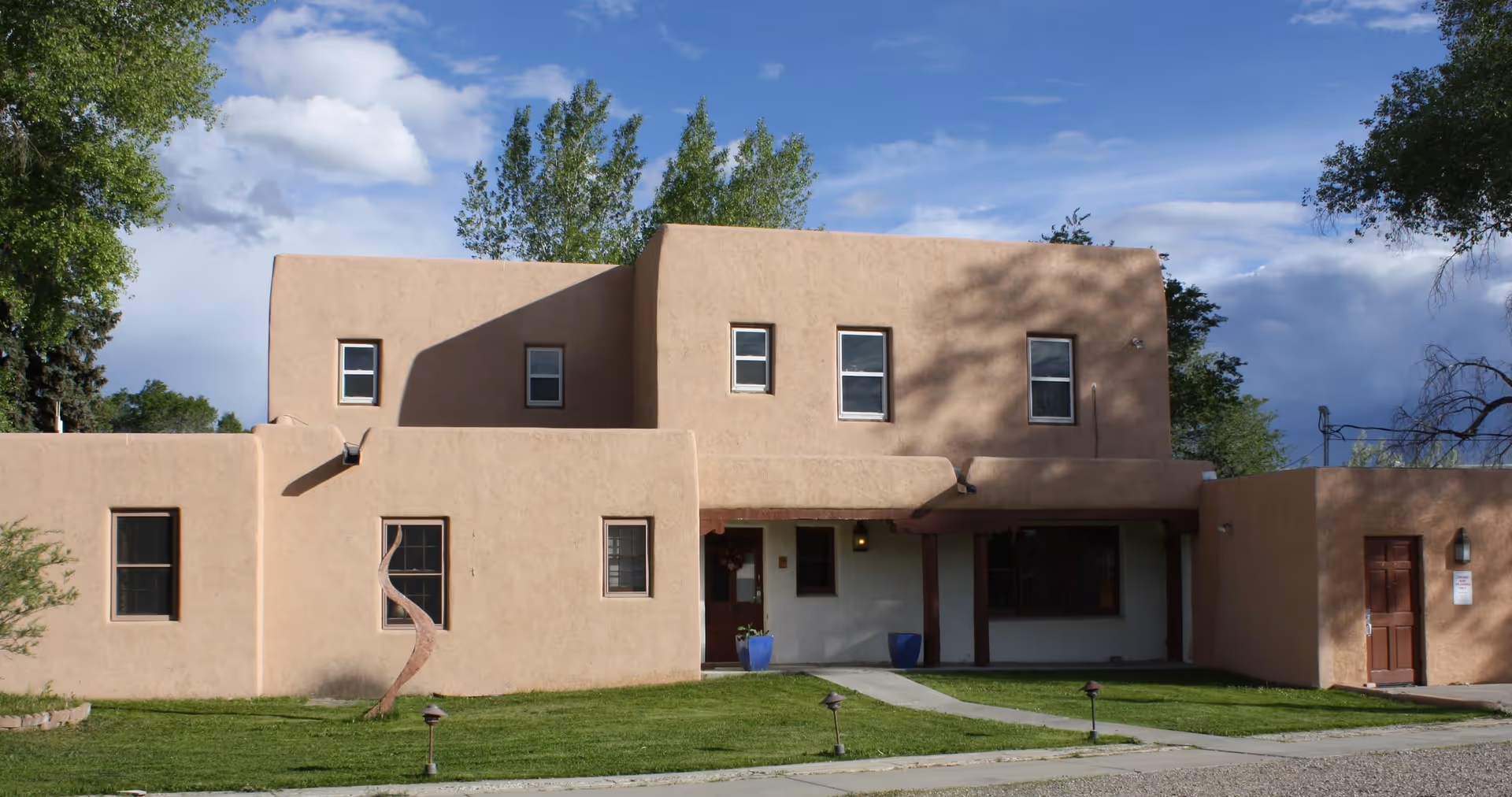 Exterior view of a two-story adobe-style building with beige stucco walls, several windows, a small covered entrance with wooden beams, green grass lawn, and a curved metal sculpture in front. Trees and a partly cloudy blue sky are visible in the background.