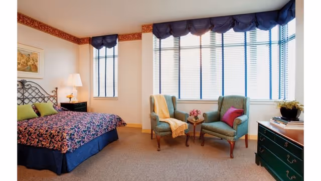 A bright bedroom with a large bed covered in a floral bedspread, two green pillows, and a dark blue bed skirt. There is a nightstand with a lamp and a small flower vase next to the bed. Two upholstered armchairs with a small table between them are positioned near a large window with blue blinds and a blue valance. A green dresser with books and a plant on top is also visible. The walls are light-colored with a decorative border near the ceiling.