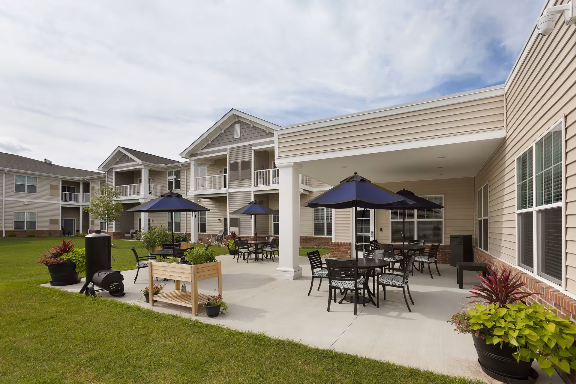Outdoor courtyard patio of a multiunit senior living building with tables, chairs, and blue umbrellas on a concrete terrace.