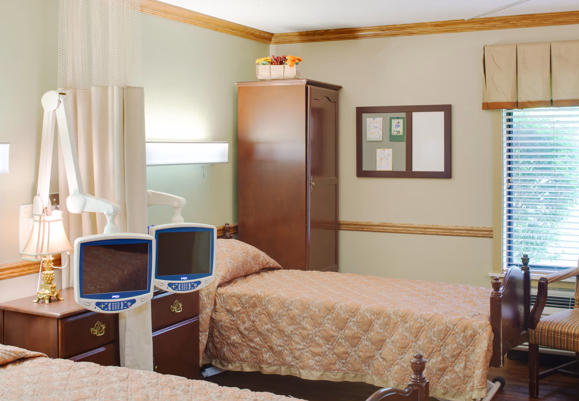 A bedroom in a senior living facility with two single beds covered in beige quilted bedspreads. Each bed has a mounted television screen at the foot. There is a wooden nightstand with a lamp between the beds, a wooden wardrobe against the wall, a bulletin board with papers pinned on it, and a window with blinds and a valance letting in natural light.