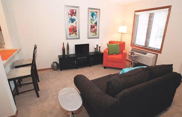 A cozy living room with a dark sofa facing a black TV stand holding a small flat screen TV, decorative bottles, and candles. There is a red armchair with a green pillow next to a window with wooden blinds and an air conditioning unit below it. A floor lamp is positioned behind the armchair, and two colorful floral paintings hang on the wall above the TV stand. Two bar stools are placed at a counter on the left side.