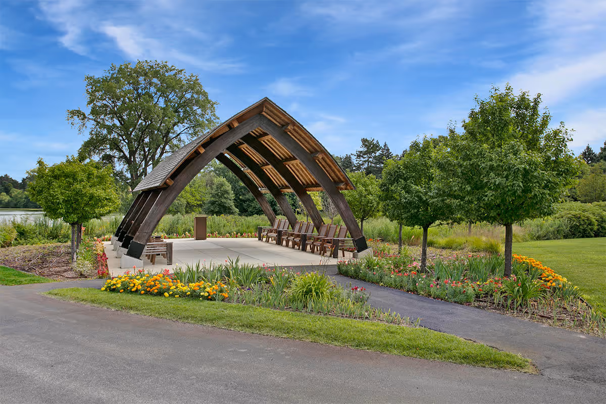 Outdoor pavilion with a wooden arched roof and several wooden chairs underneath, surrounded by green trees, colorful flower beds, and a paved pathway, set against a blue sky with some clouds.