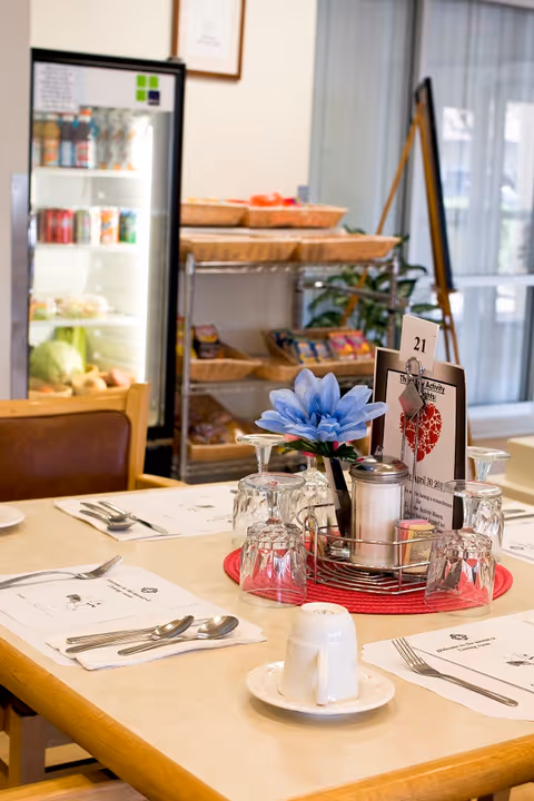 A dining table set with placemats, utensils, upside-down glasses, a small flower arrangement, and a condiment holder with sugar and salt shakers. In the background, there is a refrigerator stocked with beverages and a shelving unit with baskets containing snacks or food items.