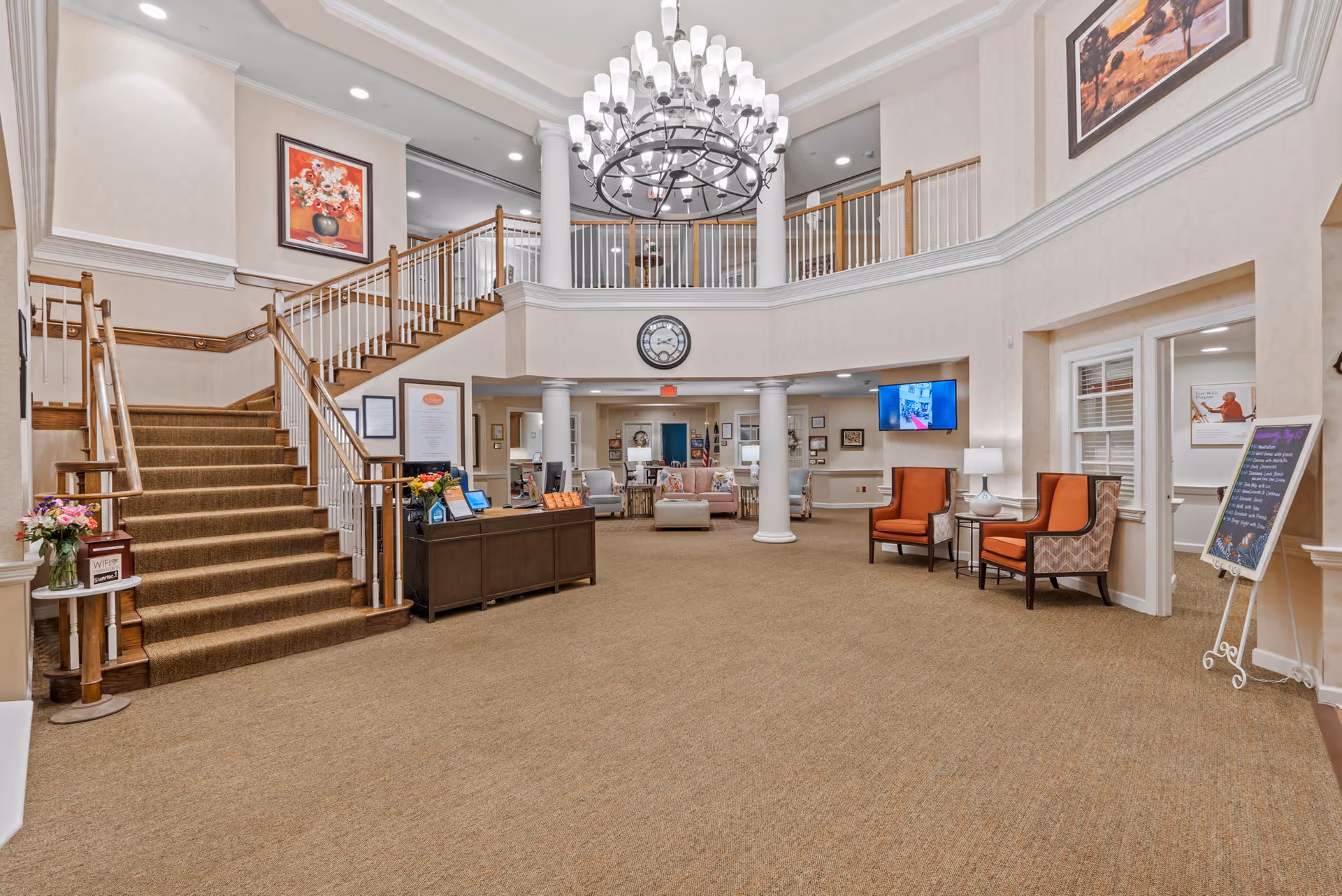 Spacious two-story senior living lobby with a central chandelier, staircase, reception desk, and seating areas.