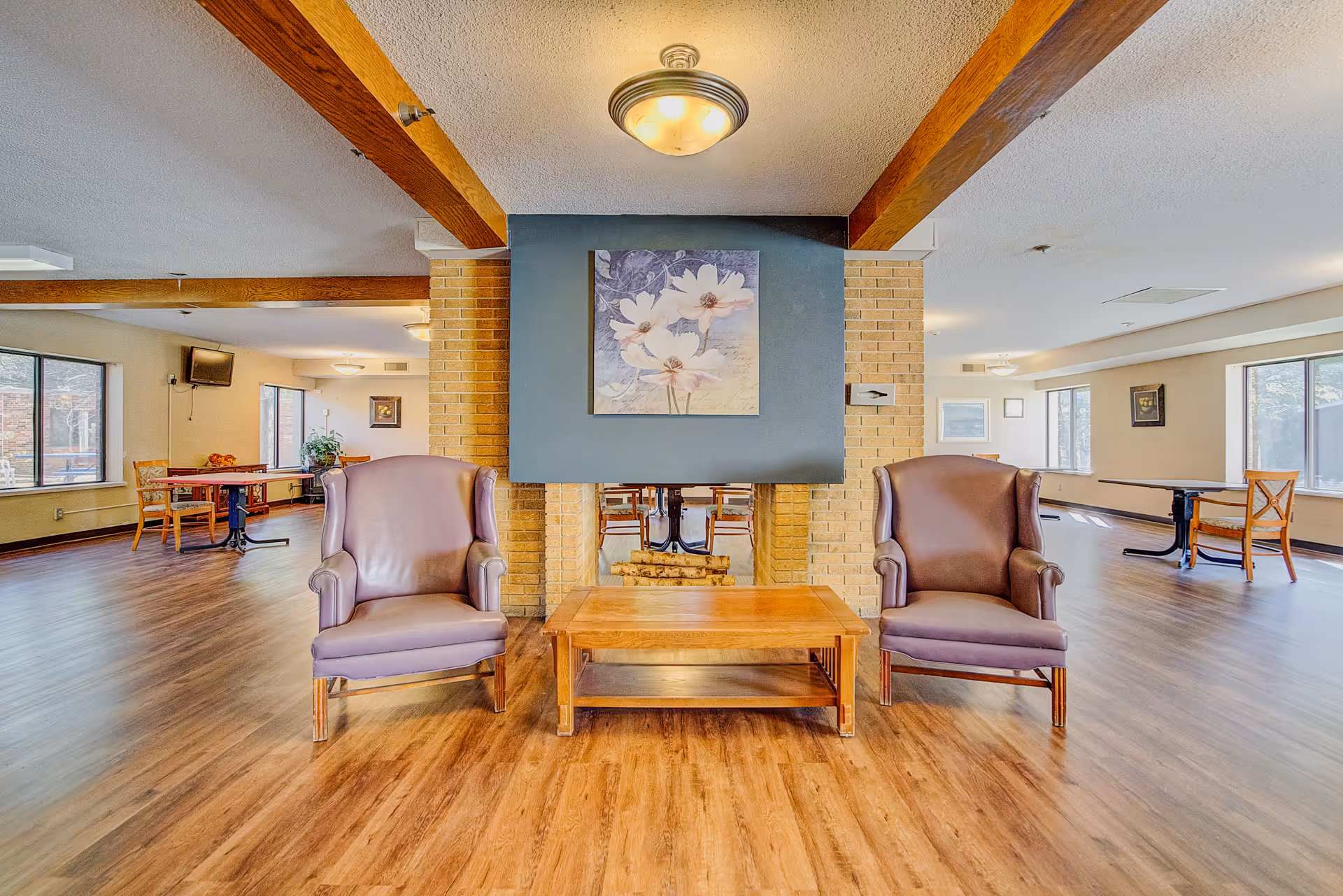 A spacious common area with wooden flooring and exposed wooden beams on the ceiling. Two purple armchairs are placed on either side of a wooden coffee table in front of a brick fireplace with a blue accent wall and a floral painting above it. In the background, there are several tables and chairs near large windows letting in natural light.