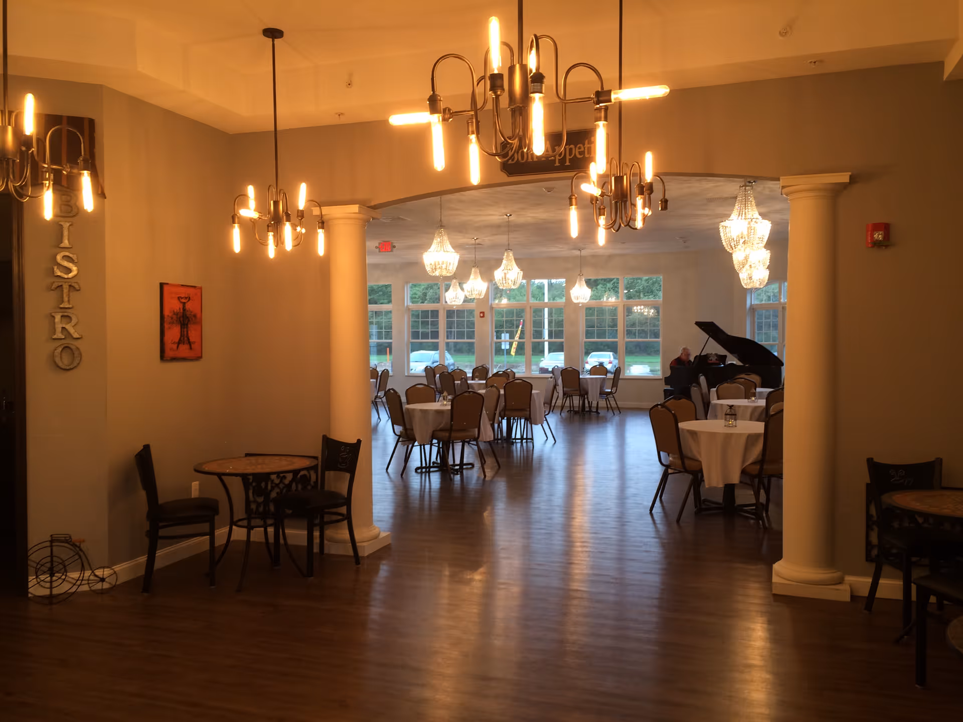 Interior view of a dining area with multiple round tables and chairs arranged neatly. The room features elegant chandeliers and modern hanging lights. A grand piano is visible in the background near large windows letting in natural light. The word 'BISTRO' is displayed vertically on a wall near the entrance.