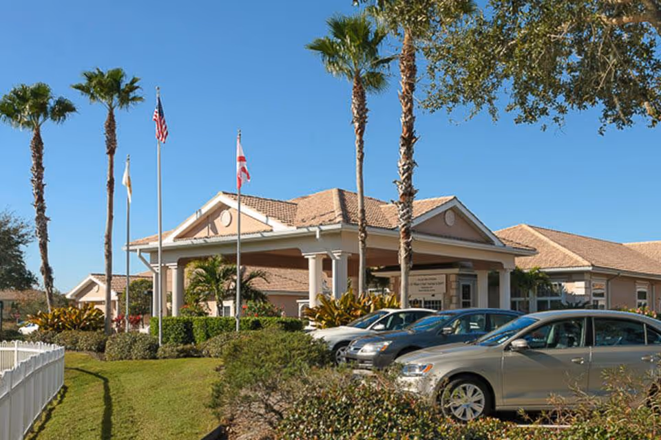 Front entrance of a senior living facility with a covered drive-through, flagpoles, palm trees and parked cars.