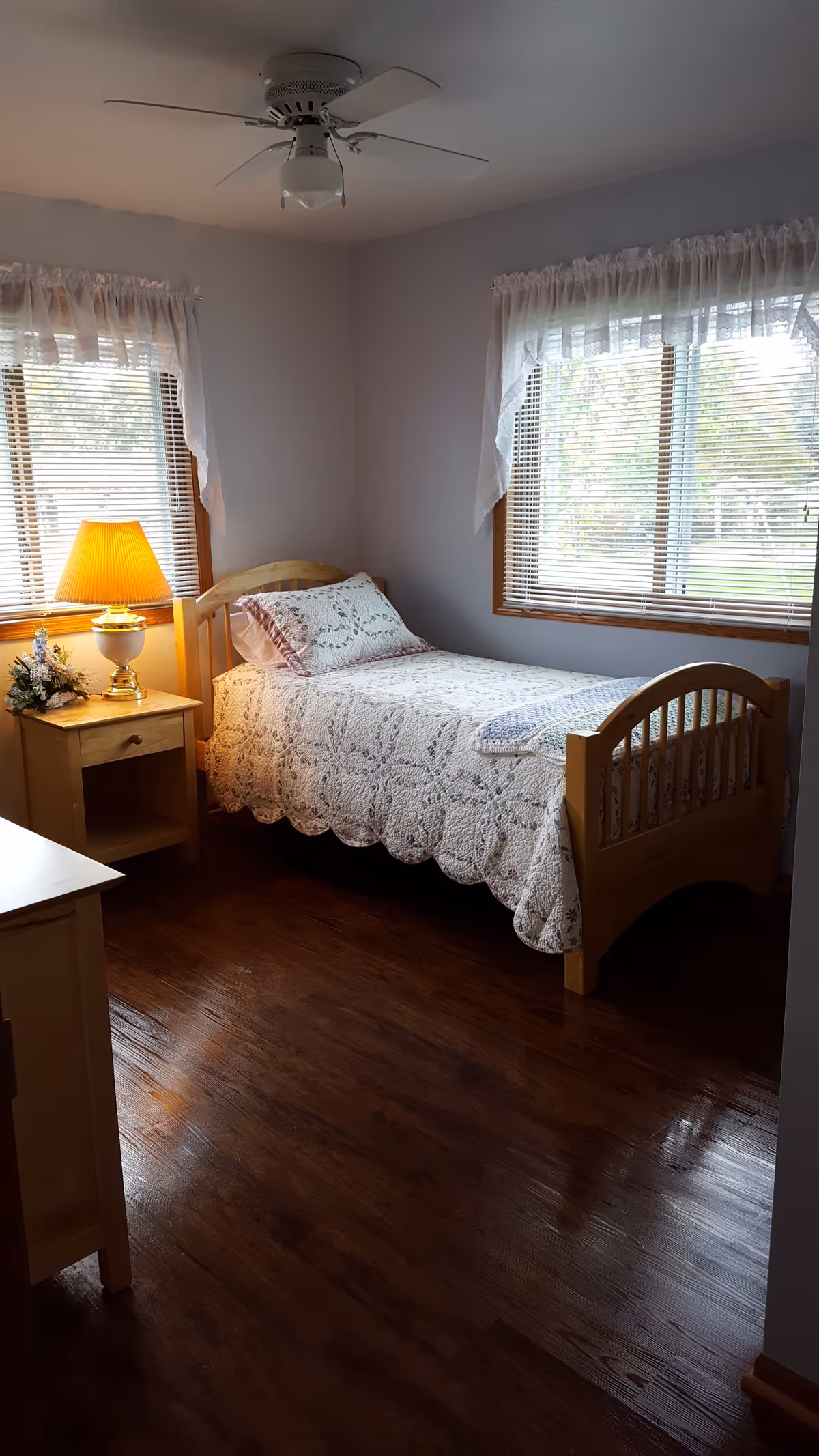 A cozy bedroom with a single wooden bed covered in a white quilted bedspread and a matching pillow. There are two windows with white lace curtains letting in natural light. A wooden nightstand beside the bed holds a lit table lamp and a small floral arrangement. The room has wooden flooring and a white ceiling fan with a light fixture.