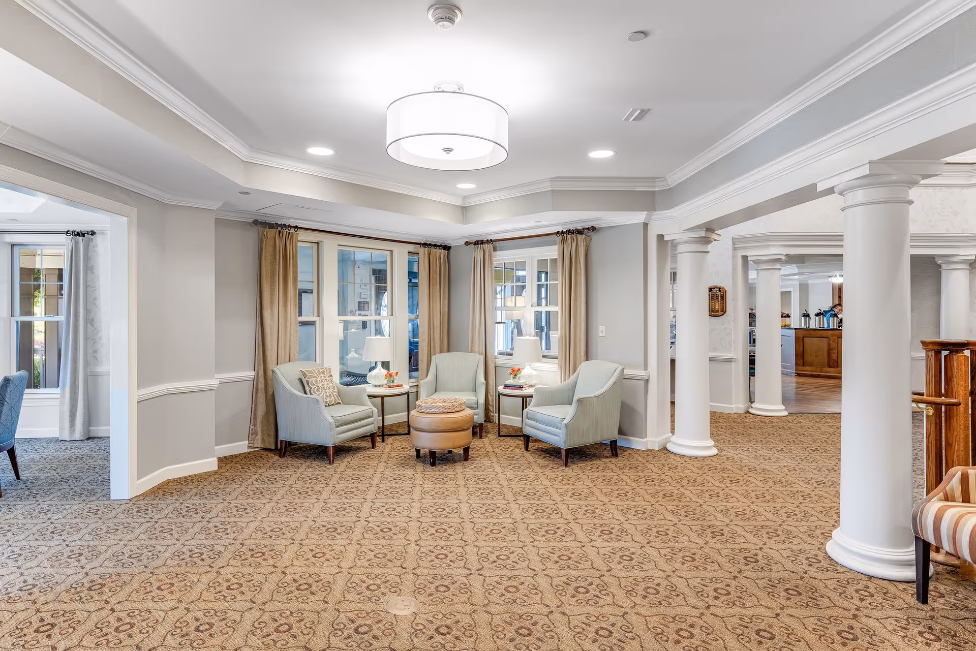 A bright and spacious common area in a senior living facility featuring three light blue armchairs arranged around a round ottoman, two side tables with lamps and small flower arrangements, beige patterned carpet, large windows with beige curtains, white columns, and soft overhead lighting.