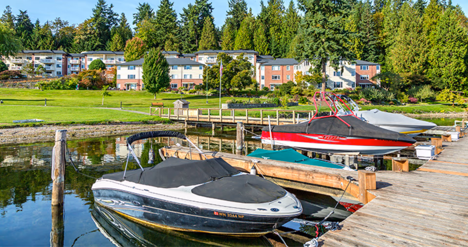 A waterfront scene at Covenant Living at the Shores showing several boats docked at a wooden pier with a grassy area and multi-story residential buildings in the background surrounded by trees.