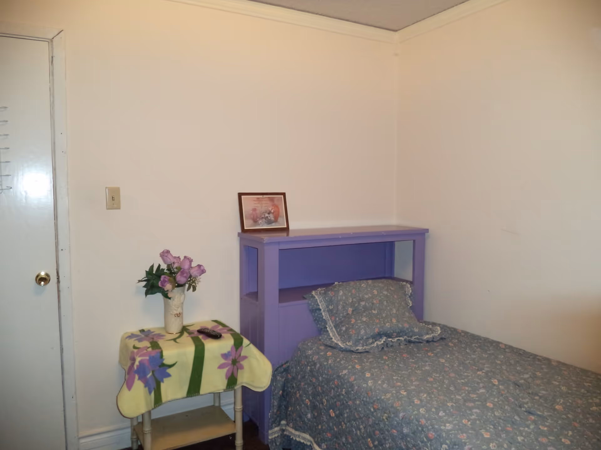 Small bedroom with a single bed featuring floral bedding, a purple headboard shelf and a side table with a vase of flowers.