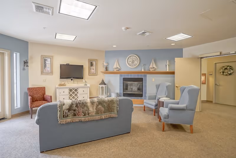 A cozy living room area with a blue sofa draped with a decorative throw, two blue armchairs, and a red armchair near a window. The room features a fireplace with nautical decorations on the mantel, a wall-mounted TV above a white cabinet, and beige carpet flooring. The walls are painted in soft blue and beige tones, and there is an open door leading to another room.