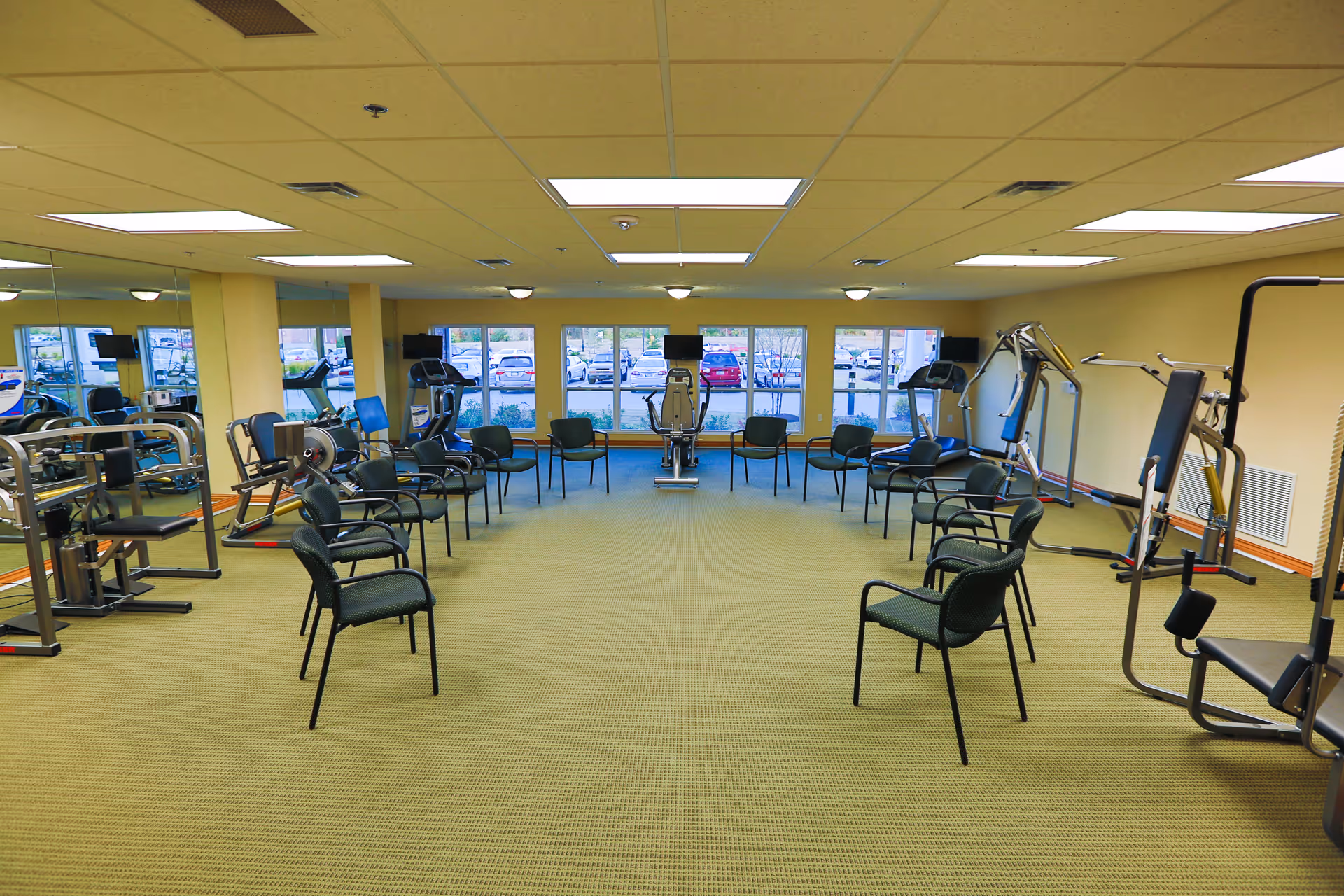 A spacious fitness room with exercise equipment along the walls and chairs arranged in a large circle in the center. The room has a beige carpet, yellow walls, large windows showing a parking lot outside, and a ceiling with fluorescent lights.