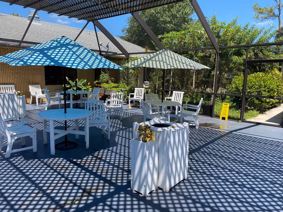 A screened outdoor patio with white tables and chairs, large umbrellas, planters, and lattice shadows on the floor.
