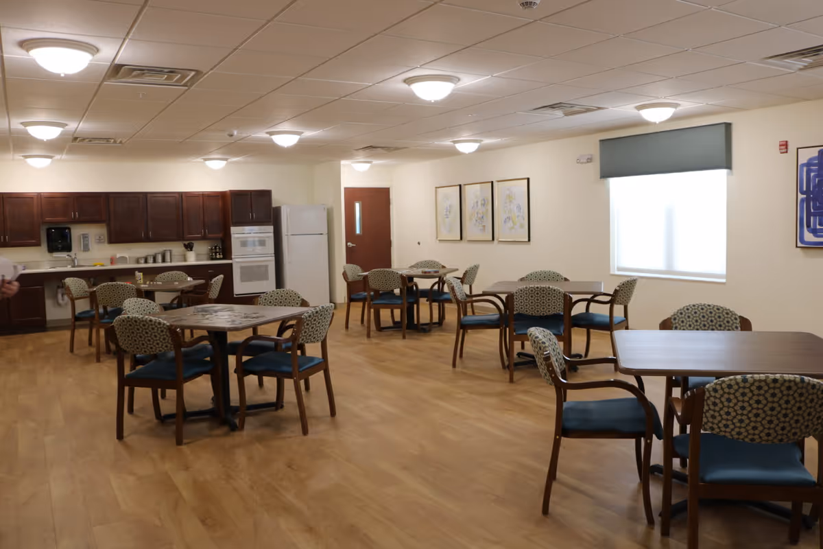 Spacious senior facility dining area with multiple tables and chairs and a kitchenette along the back wall.