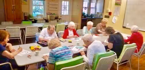 A group of elderly people seated around a rectangular table in a well-lit room, engaged in drawing or coloring activities with papers and colored pencils in front of them.