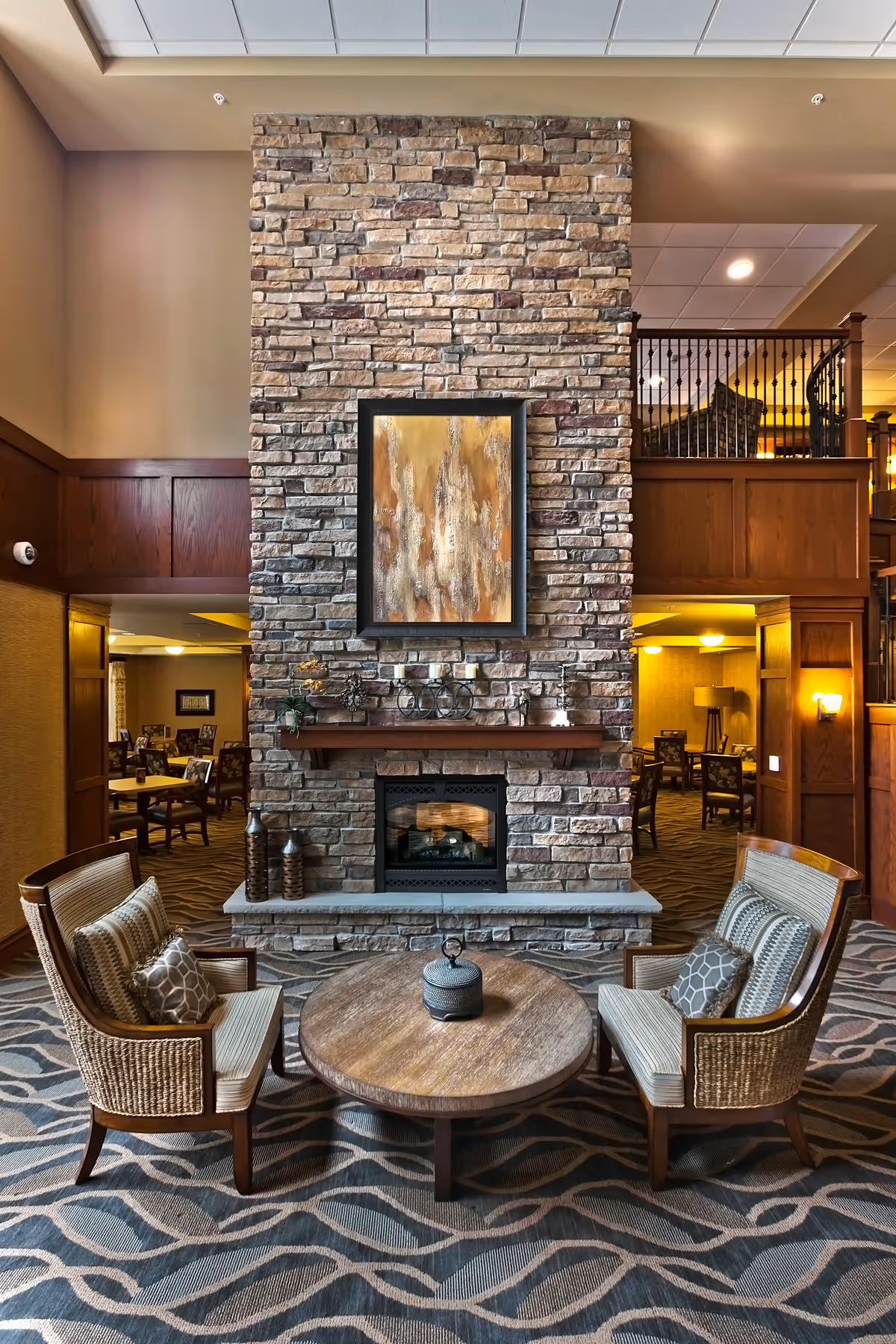 Cozy seating area with two cushioned armchairs facing a round wooden coffee table, set in front of a tall stone fireplace with a wooden mantel and abstract artwork above it. The room features warm lighting, wood paneling, and a patterned carpet, with a dining area visible in the background.