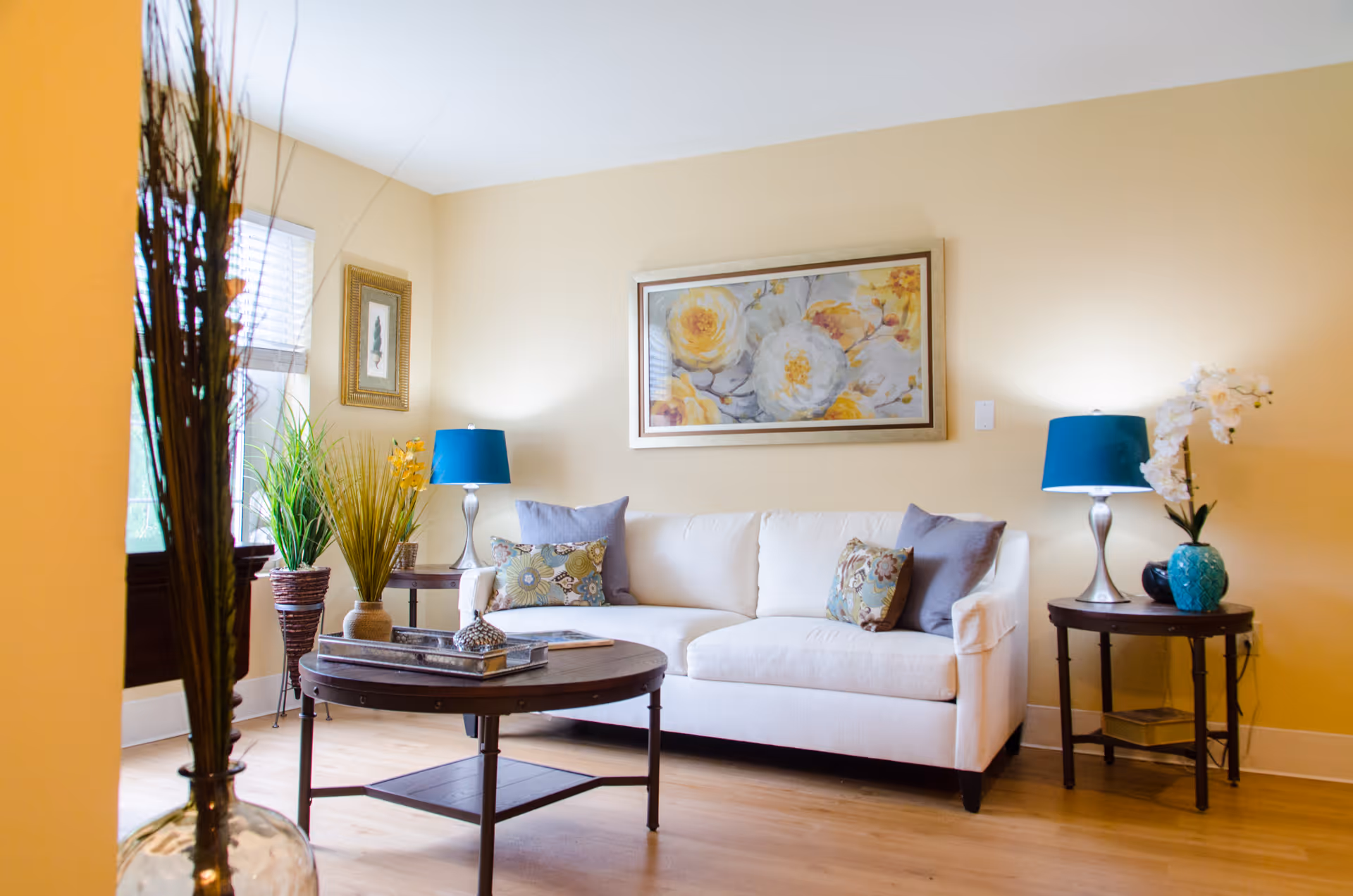 A bright living room with a white sofa adorned with patterned and solid pillows, two side tables each with a blue lamp, a round wooden coffee table with decorative items, potted plants near a window, and a large floral painting on the beige wall.