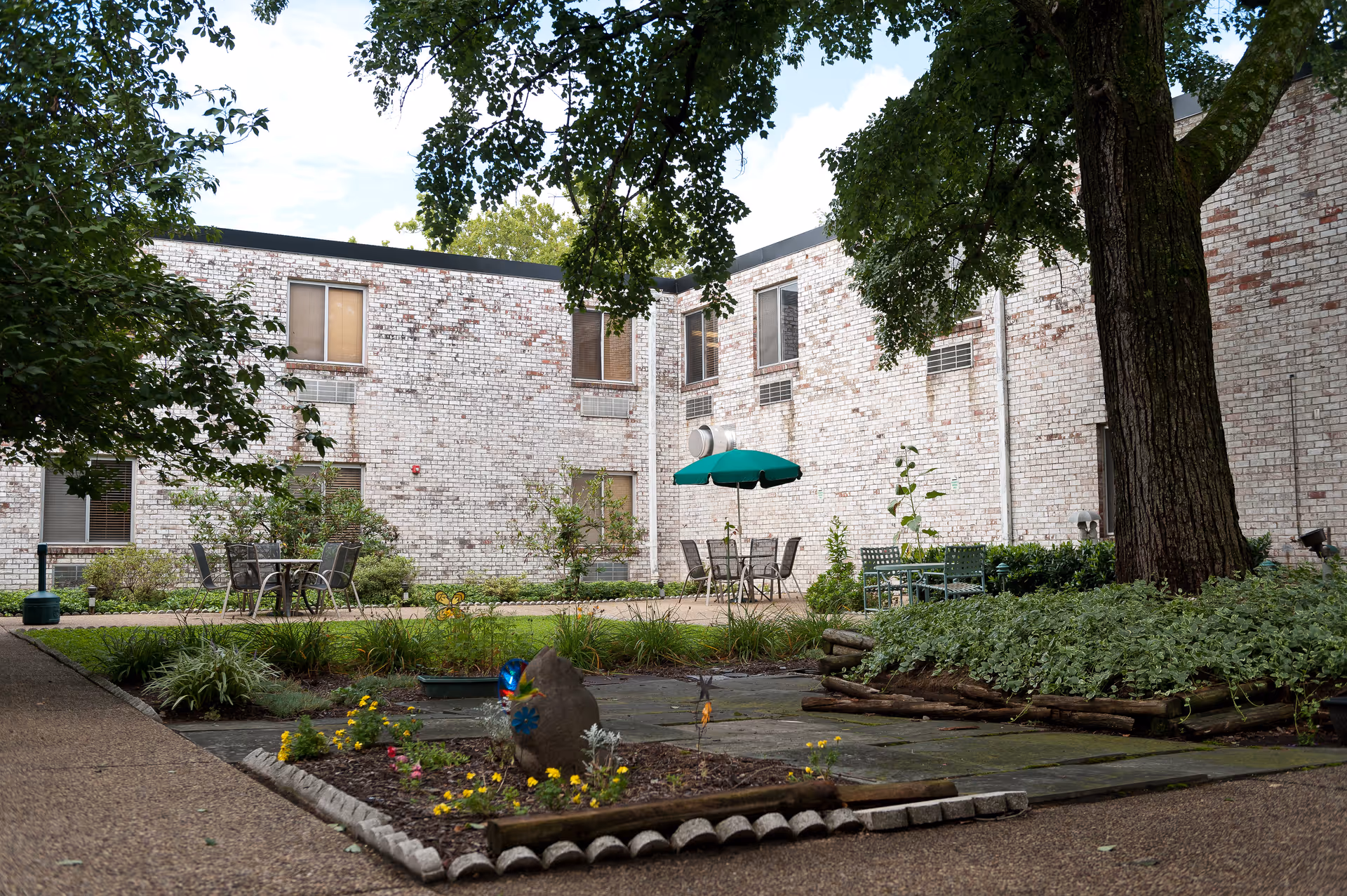 Outdoor courtyard area at Autumn Lake Healthcare at Pikesville featuring a garden with flowers, a large tree, patio tables and chairs, and a green umbrella against a backdrop of a white brick building.