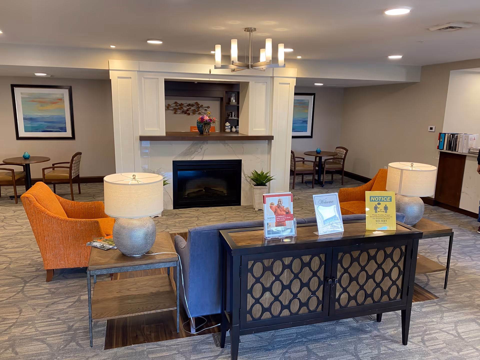 A cozy senior living facility common area with two orange armchairs and a blue sofa arranged around a fireplace. There are two table lamps on side tables, a chandelier overhead, and framed artwork on the walls. Small tables with chairs are visible in the background. On the back of the sofa, there are informational signs including a welcome sign and a social distancing notice.
