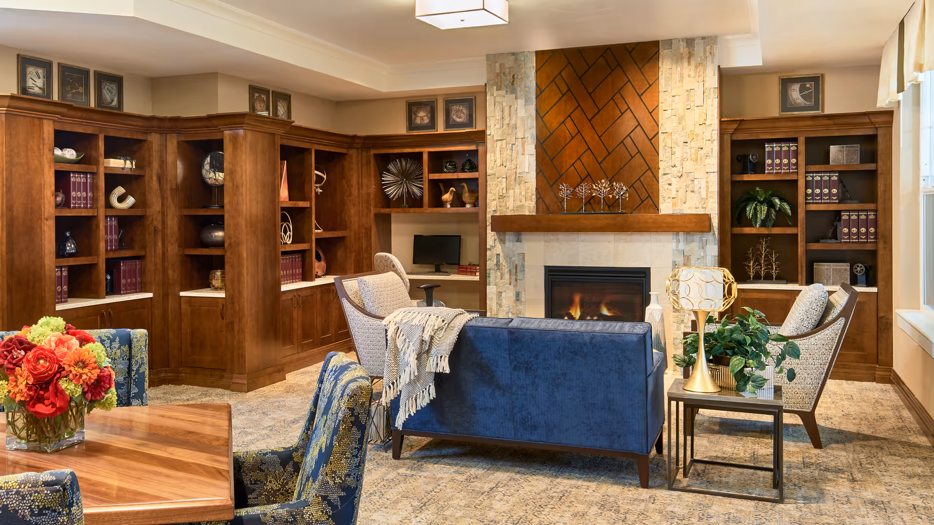 A cozy living room area with a lit fireplace surrounded by stone and wood paneling. The room features built-in wooden bookshelves filled with decorative items and books. There is a blue upholstered sofa, two patterned armchairs, a side table with a lamp and a plant, and a wooden table with floral arrangements. The space is warmly lit and inviting.