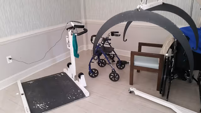 A room with medical and mobility equipment including a white patient lift, a blue walker, a wooden chair with light blue cushion, and a wheelchair partially visible on the right side. The room has light-colored walls and flooring.