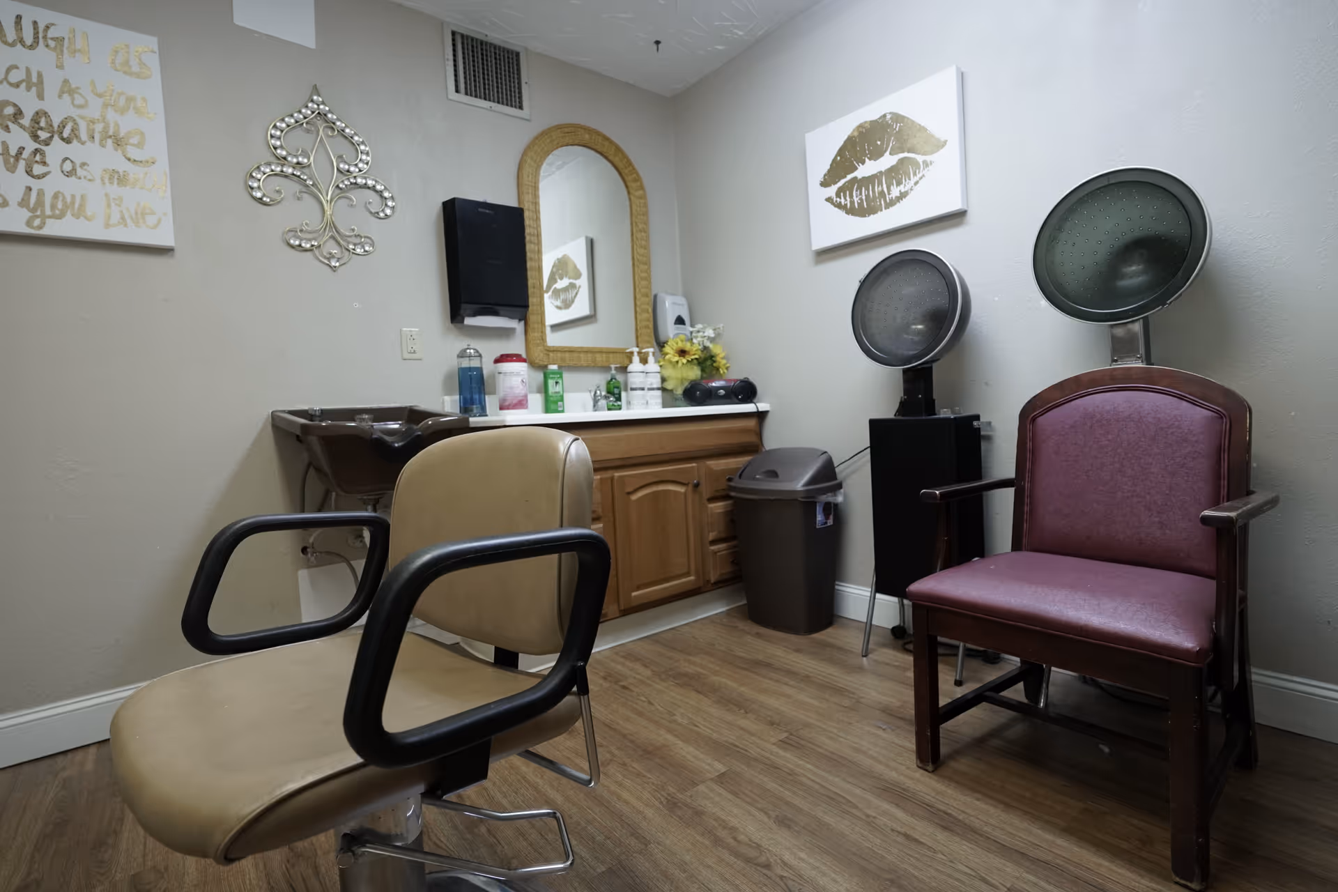 Interior of a small salon room with a beige salon chair in the foreground and a maroon salon chair with a hair dryer hood in the background. There is a wooden cabinet with a mirror above it, various hair care products on the counter, a trash bin, and wall decorations including a gold lip print and a decorative metal piece.