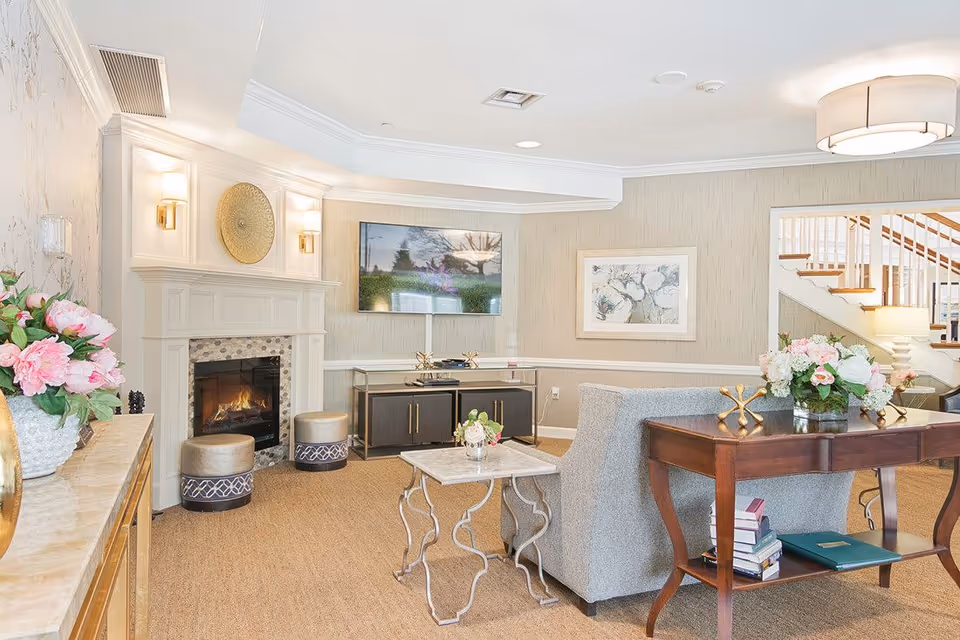 A bright and cozy living room area with a lit fireplace, a mounted flat-screen TV, a gray sofa, a wooden console table with books and floral arrangements, and a small decorative side table. The room features beige walls, carpeted floor, and a staircase visible in the background.