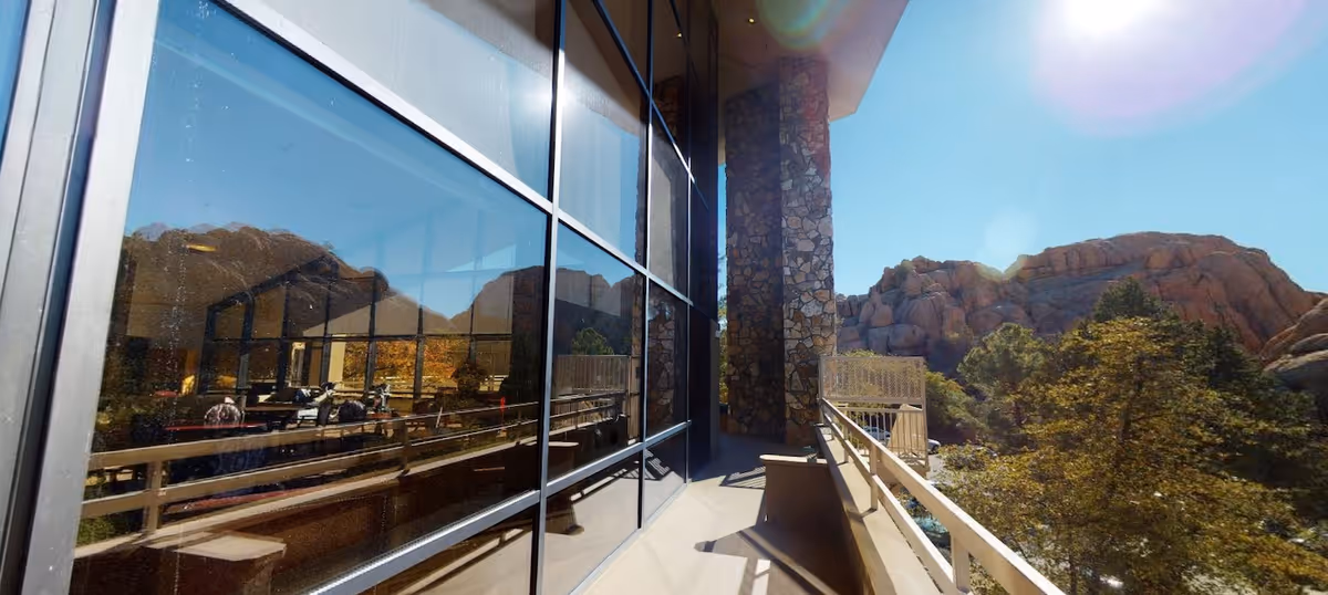 A sunny outdoor balcony area with a stone pillar and railing, overlooking trees and rocky hills in the background. The glass windows on the left reflect the outdoor scenery and some indoor furniture.