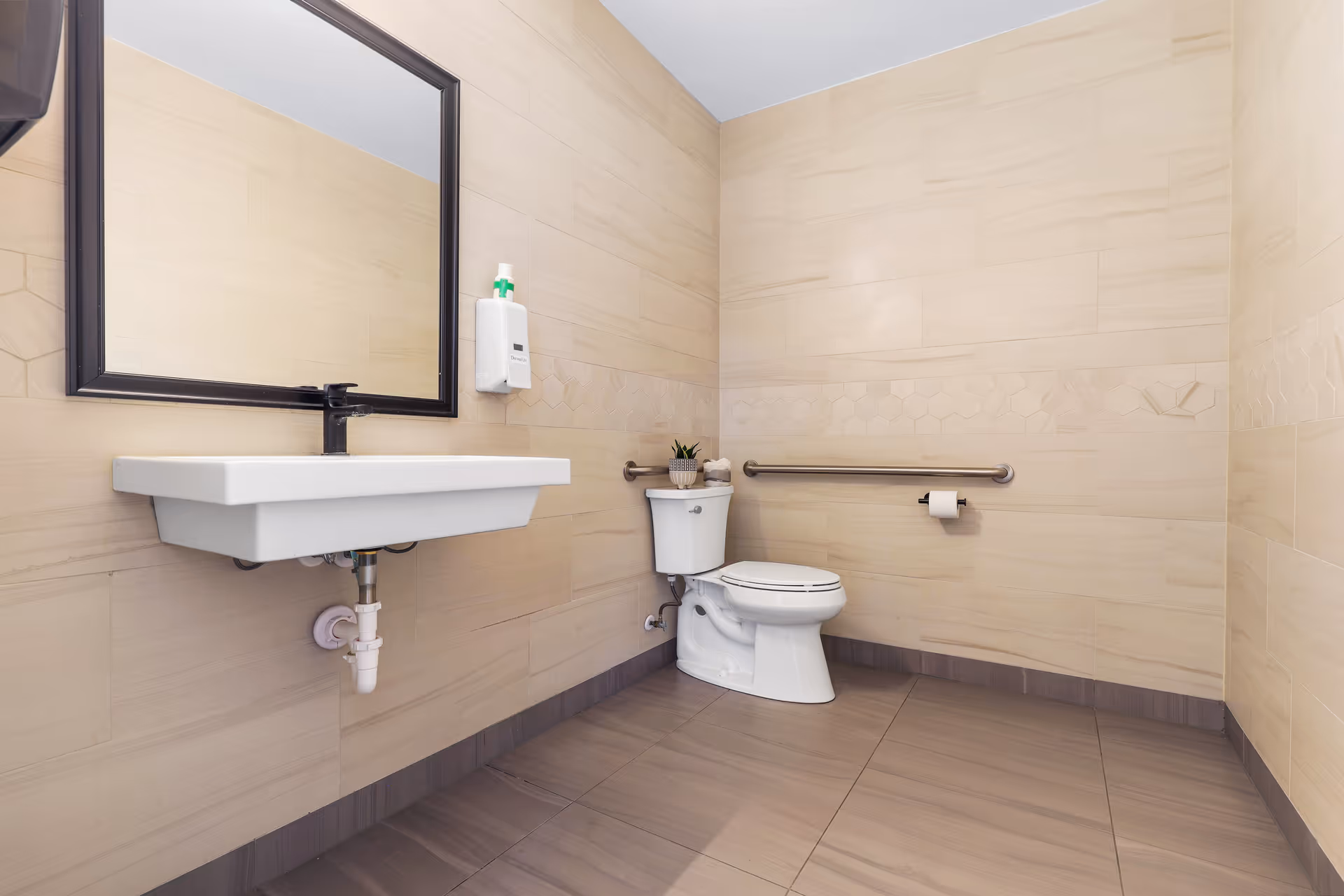 Accessible restroom with a wall-mounted sink and mirror, toilet with grab bar, and soap dispenser.