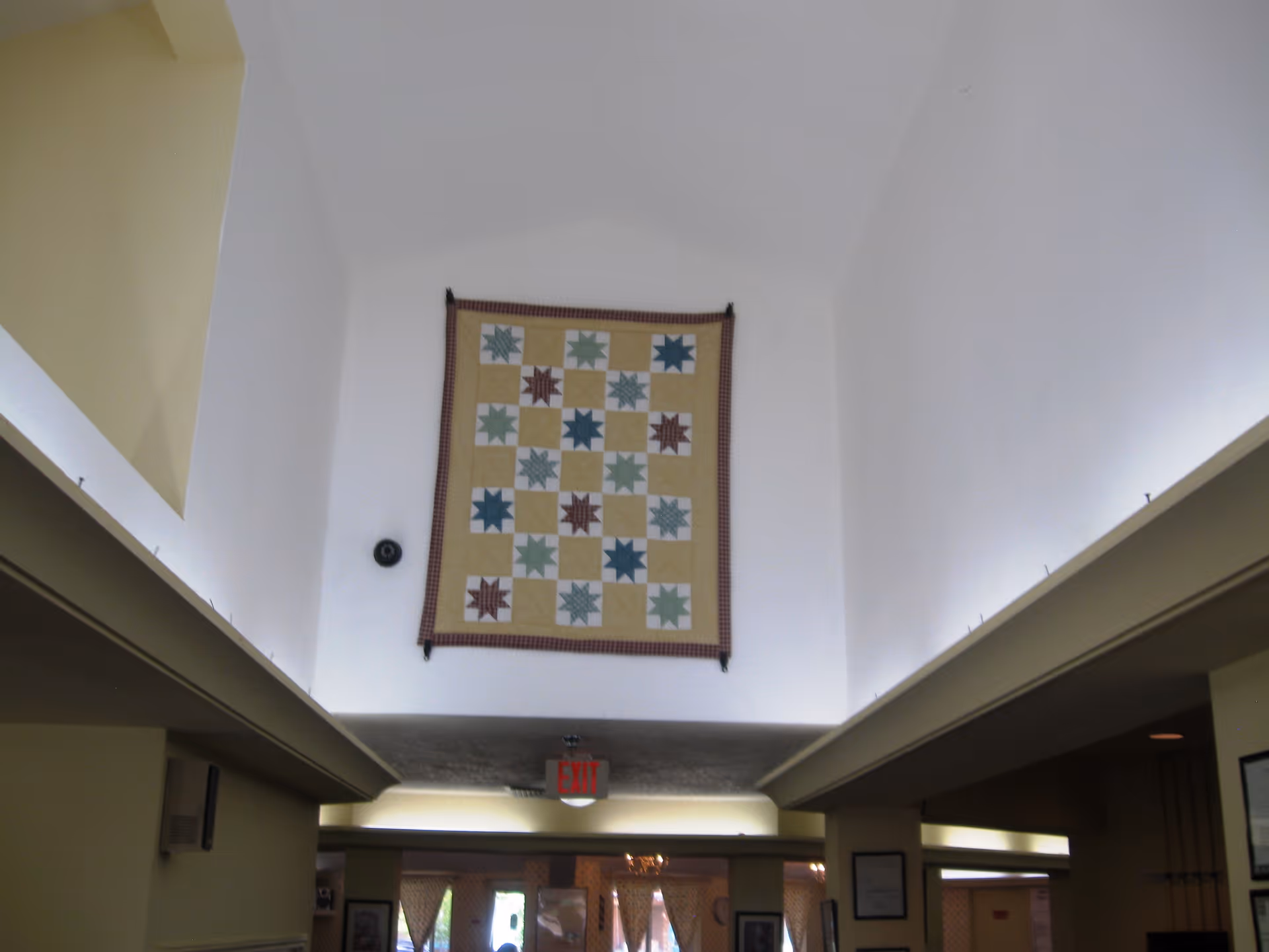A quilted wall hanging displayed high above an interior hallway entrance with an EXIT sign below.