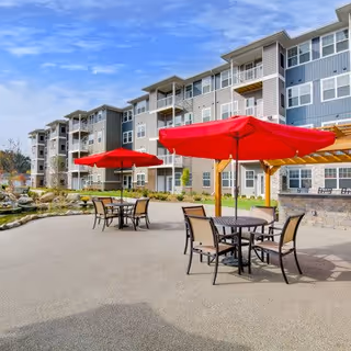 Outdoor patio area with tables and chairs shaded by large red umbrellas in front of a multi-story residential building with balconies under a partly cloudy sky.