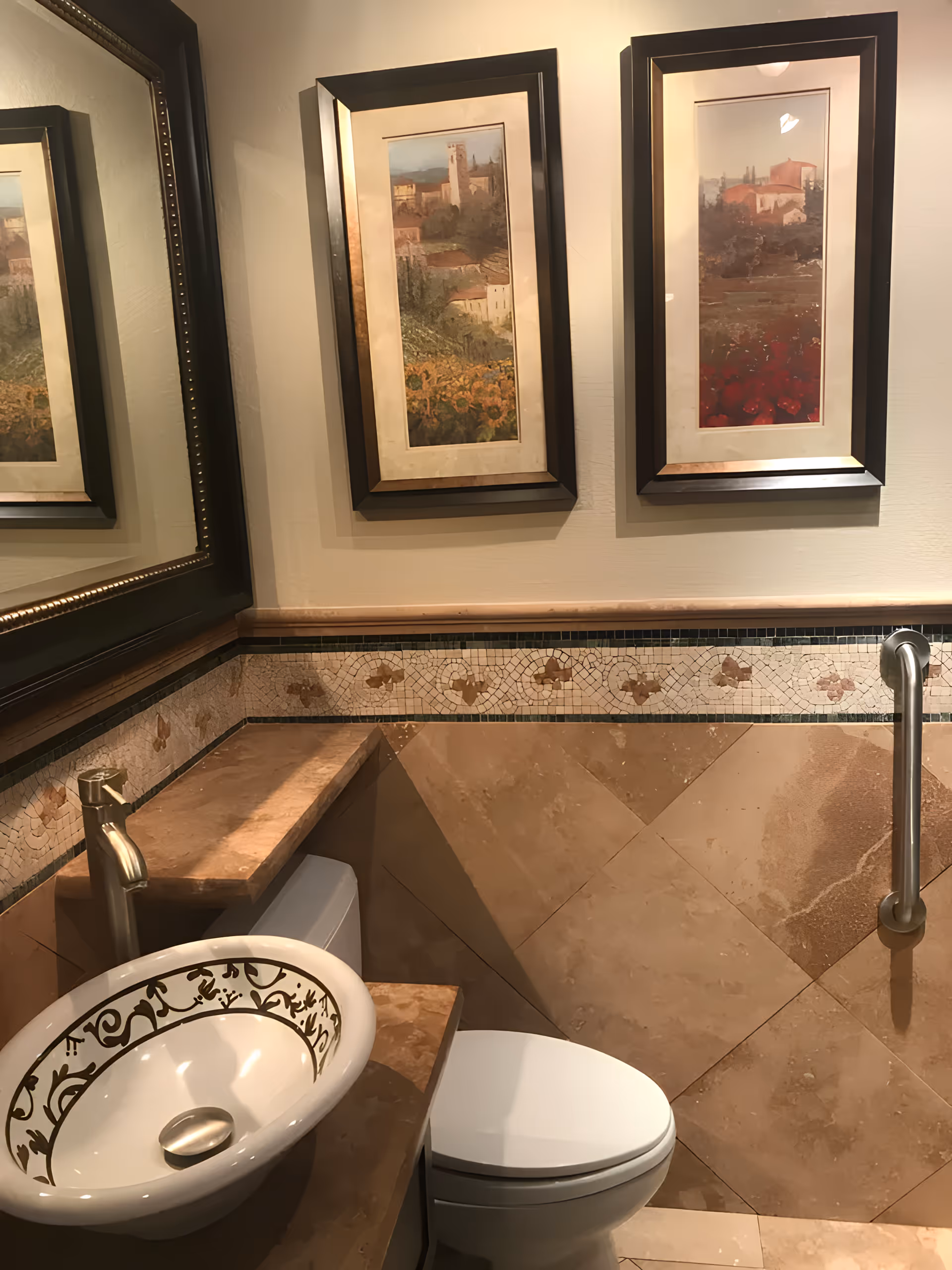 A small bathroom with a decorative white sink featuring a gold floral pattern, a modern faucet, a white toilet, beige tiled walls with a mosaic border, a grab bar on the right wall, and two framed landscape paintings above the toilet.