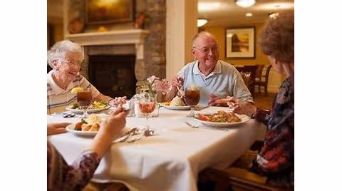 Four elderly people sitting around a dining table in a cozy room, enjoying a meal together with plates of food and glasses of iced tea, with a fireplace and framed pictures in the background.