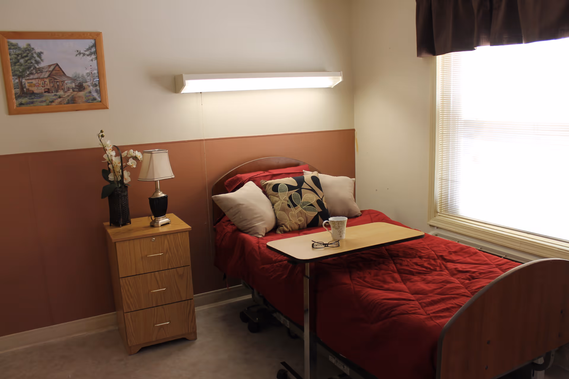 A small bedroom in a healthcare facility with a single bed covered in a red quilt and several pillows. A wooden bedside table with a lamp and a vase with flowers is next to the bed. Above the bed is a wall-mounted light fixture. A window with blinds and a dark valance lets in natural light. A framed painting hangs on the wall.