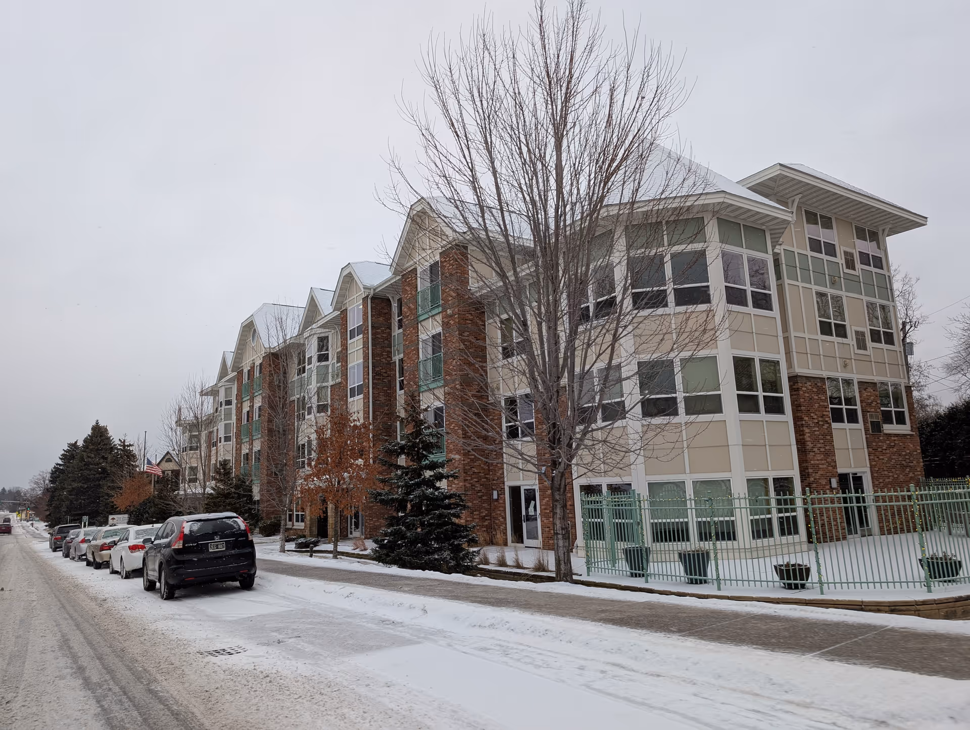 Front exterior of a multi-story senior living building beside a snowy street with parked cars and bare trees.
